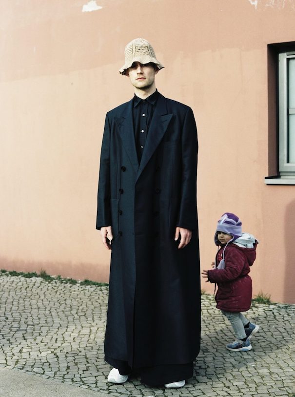 Andra Dumitrascu Long black coat beige hat orange wall kid